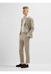 Bežni teksturirani suit s slim-fit hlačama i belom košuljom. Model nosi tamnosmeđe cipele na lofer s zlatnim detaljima i belim čarapama.