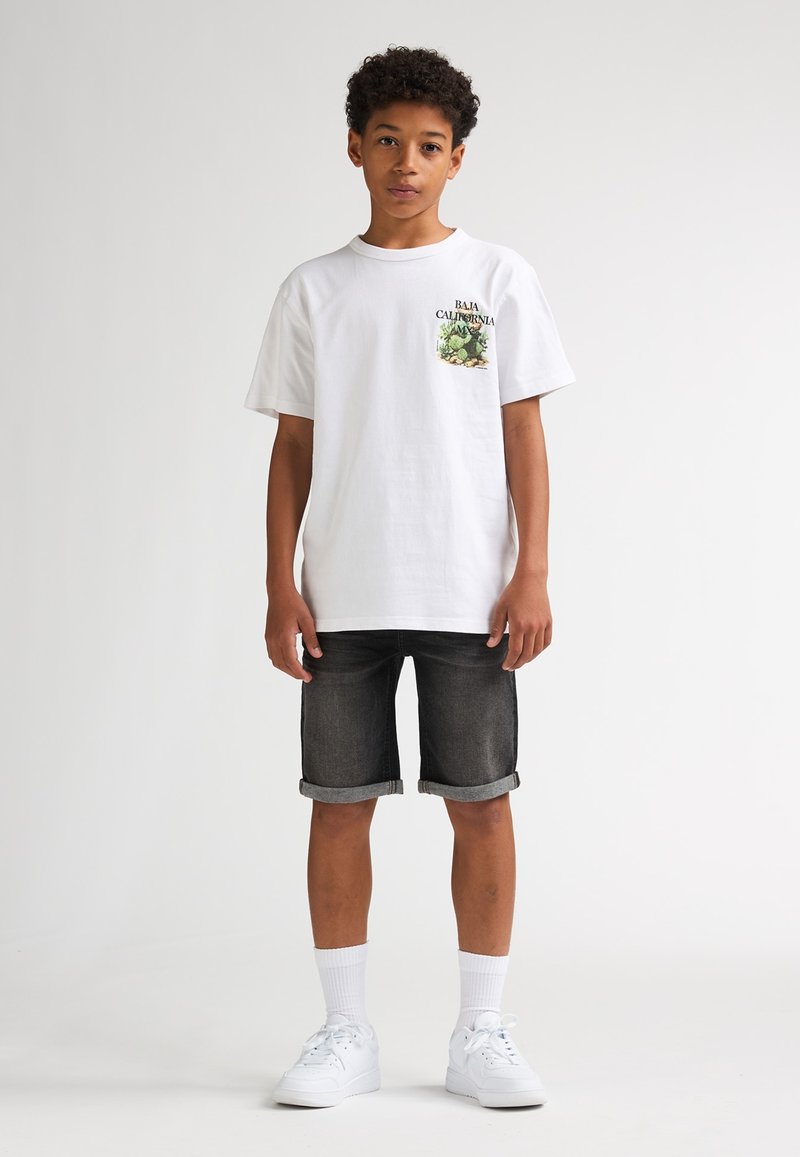 T-shirt blanc en coton avec un motif graphique sur la poche indiquant "Baja California", associé à un short en jean foncé et des baskets blanches.