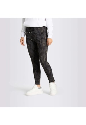 Personne portant un pantalon de jogging noir à motif floral et des baskets blanches, debout contre un fond clair uni.