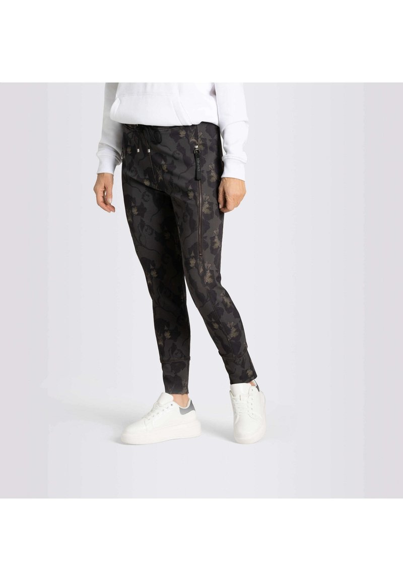Personne portant un pantalon de jogging noir à motif floral et des baskets blanches, debout contre un fond clair uni.