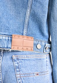 Džínová bunda svetlomodrej farby s hnedou štítkom s vyrazeným textom "TOMMY JEANS" a striebornými gombíkmi.