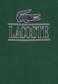 Grön texturerad tyg med en framträdande marinblå Lacoste-logotyp med ett krokodilmotiv och horisontella ränder.