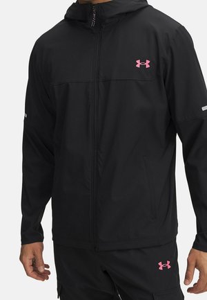 Czarna kurtka z zamkiem i spodnie z różowymi logo Under Armour noszone przez osobę, pokazujące kieszenie kurtki i odblaskowe detale na rękawach.