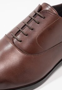 Chaussure en cuir marron avec une texture lisse, dotée d'un bout arrondi, de six œillets et de lacets d'une teinte marron plus foncée.