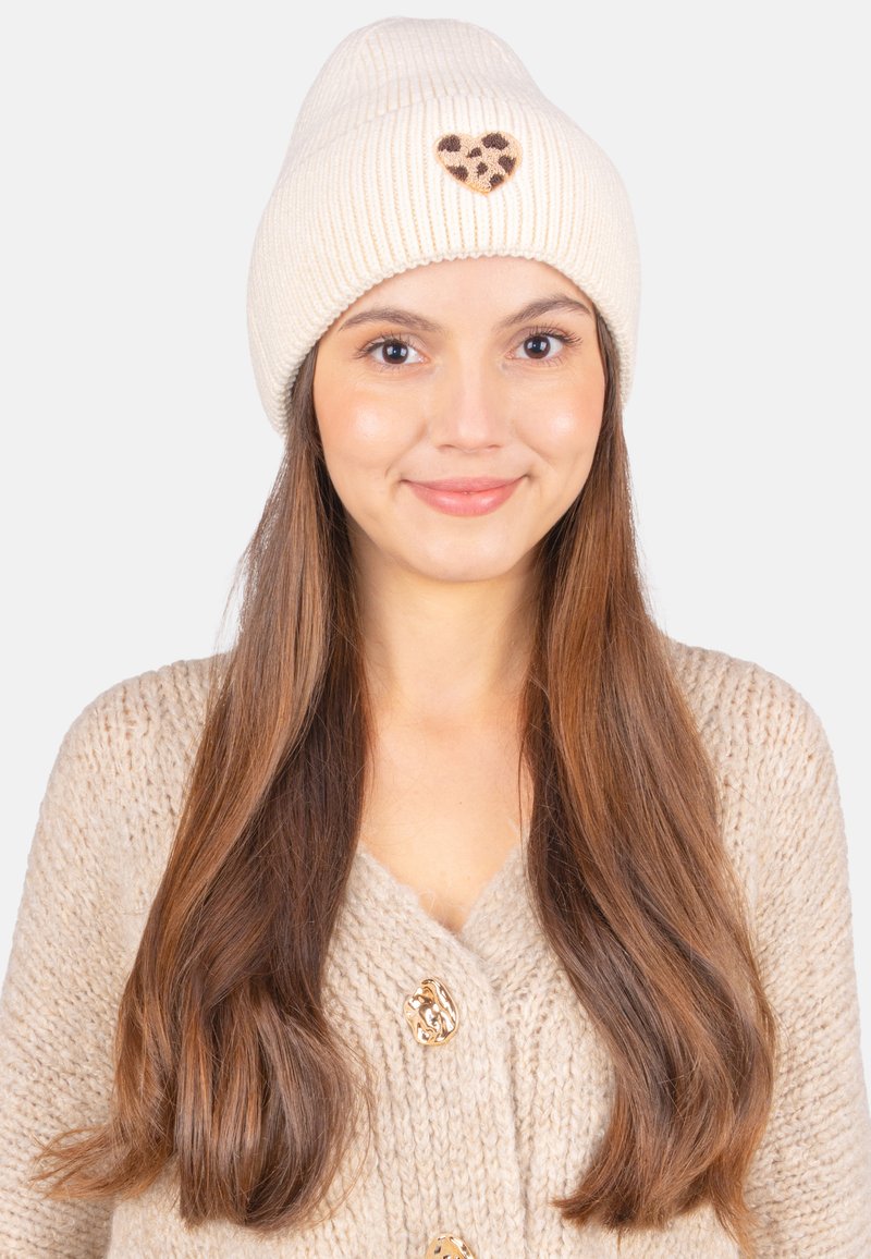 Bonnet en tricot côtelé crème avec un accent en forme de cœur en imprimé léopard. Texture douce et design ajusté, stylé avec des cheveux longs et flottants.