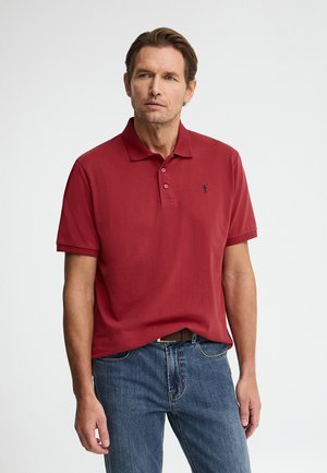 Hombre de piel clara y cabello castaño, vistiendo una camisa polo roja de manga corta y jeans azules, con un fondo blanco liso.