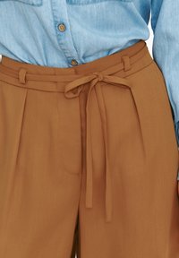 Brązowe spodnie w kant z paskiem, wykonane z gładkiego materiału. Posiadają kieszenie boczne i widoczne detale szycia.