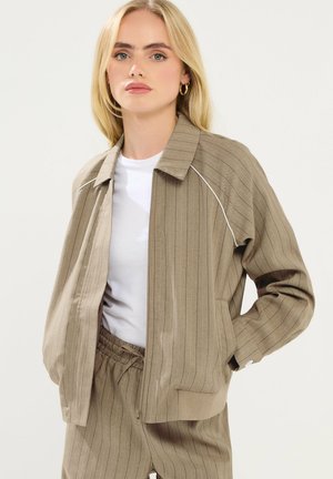 Blondynka ubrana w beżową marynarkę w prążki i pasujące spodnie na białej koszuli, stojąca z rękami w kieszeniach na jednolitym tle.