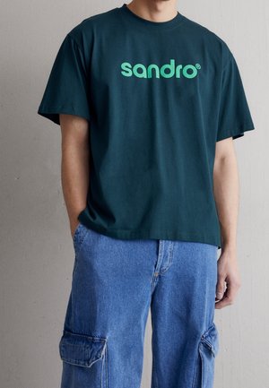 Person trägt ein dunkelgrünes T-Shirt mit "sandro"-Logo und blaue Jeans-Cargohose, steht vor einer schlichten hellgrauen Wand.