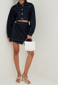 Denim crop-jakki hopeanvärisillä nappulalla, yhdistettynä samasta denimista valmistettuun hameeseen, jossa on epäsymmetrinen helma. Valkoinen käsilaukku ja vaaleanbeige korkokenkä.