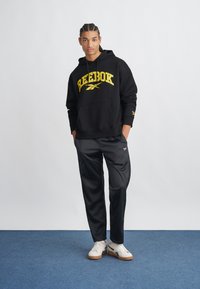 Čierna mikina s kapucňou so žltým logom "Reebok", v kombinácii s čiernymi športovými nohavicami a bielymi teniskami s gumovou podrážkou. Hladký materiál, voľný strih.