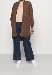 Abrigo largo marrón acolchado con puños acanalados y bolsillos, combinado con una camisa rosa claro y vaqueros oscuros de pierna ancha, terminado con zapatillas blancas.