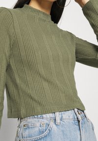 Top de manga larga de color verde oliva con cuello alto, con rayas verticales y una cintura ajustada. Combinado con unos jeans de cintura alta azules.