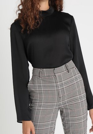Kobieta ubrana w czarną bluzkę z długim rękawem włożoną do wysokich, czarno-białych spodni w kratę na jednolitym tle.