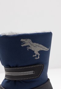 Botte en tissu bleu avec doublure en polaire blanche, présentant un motif de dinosaure gris et une sangle d'accent noire avec des coutures grises. Semelle épaisse noire.