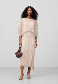 Helle rosa, plissierte Midi-Kleid mit einem cropped, strukturierten Overlay. Das Model hält eine dunkle geflochtene Tasche und trägt offene, beige Absätze.