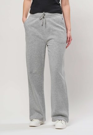 Persoon draagt lichtgrijze wijdvallende joggingbroek, witte sneakers en een zwart mouwloos topje, staat met handen in de zakken.