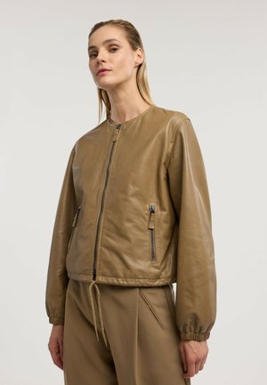 Frau trägt eine hellbraune Lederjacke mit Reißverschluss und elastischen Bündchen sowie passende hellbraune Hose vor einem schlichten hellen Hintergrund.