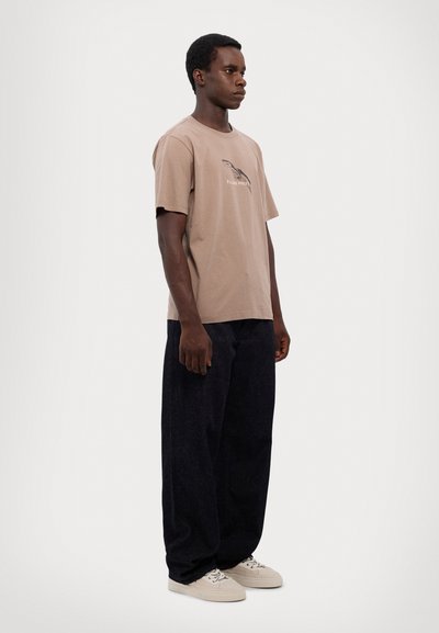Ein Mann steht, trägt ein braunes Grafik-T-Shirt, lockere dunkle Hose und hellbeige Turnschuhe, vor einem schlichten weißen Hintergrund.