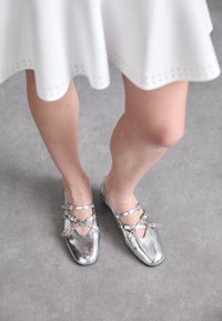 Ballerine metalliche argento con fasce incrociate, punta rettangolare e superficie liscia. La scarpa presenta una suola piatta e dettagli minimi.