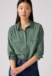 Groene button-up shirt met twee borstzakken, opgerolde mouwen en een subtiele textuur, gecombineerd met donkerdenim jeans. Minimale merklogo zichtbaar.