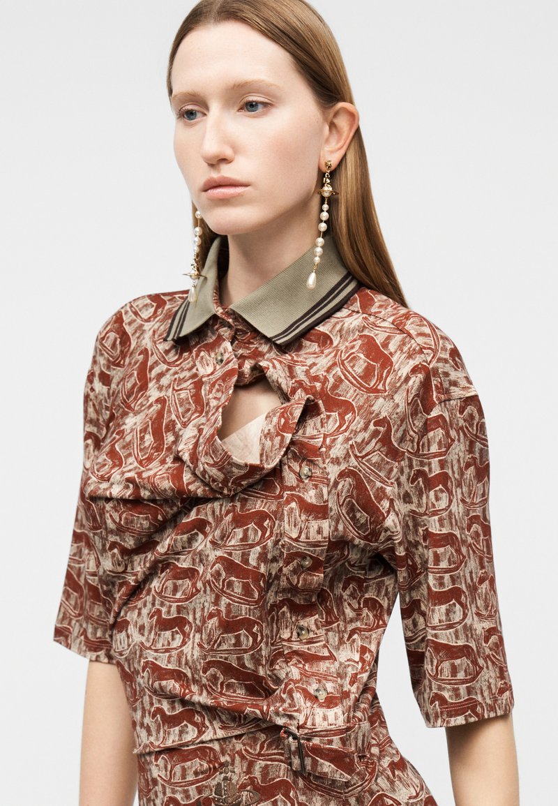 Giovane donna con lunghi capelli che indossa una camicia a fantasia rustica e beige con bottoni, colletto verde e lunghi orecchini a goccia di perle.