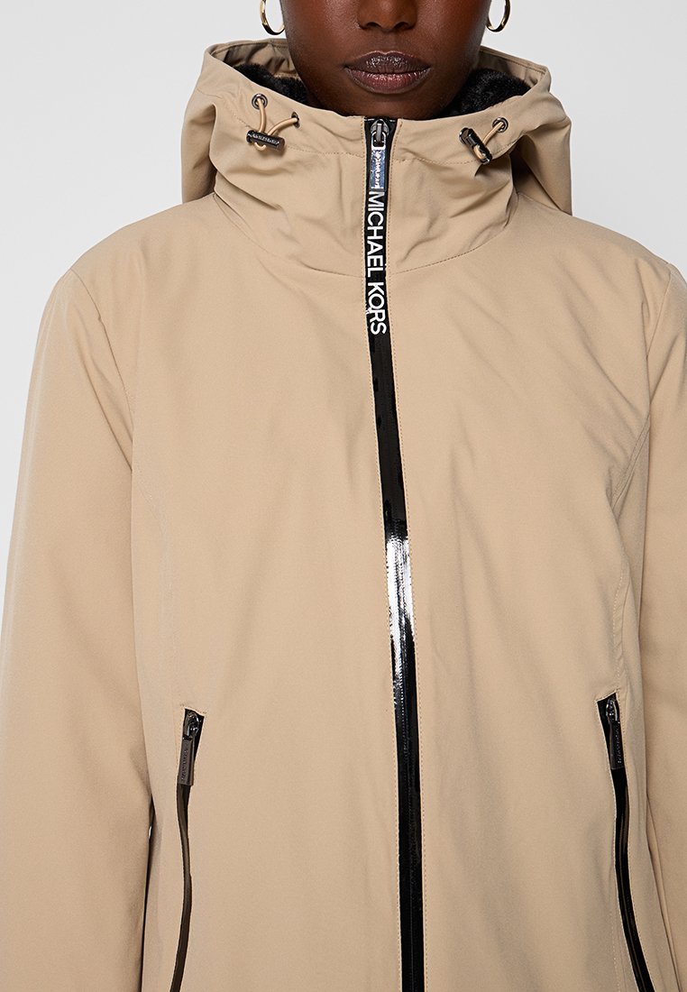 Beige gewatteerde jas met een zwarte rits en branding, voorzien van slanke zijzakken en een zachte textuur.