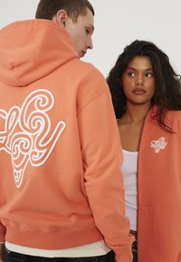 Oransje hettegenser med hvit logo-detalj på baksiden og forsiden. Har ribbestrikkede mansjetter og kant, avslappet passform og myk tekstur.