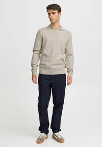 Beige pulóver V nyakkal és gallérral, puha anyagból készült. Sötét farmernadrággal és fehér-tan dizájnnal ellátott sportcipőkkel párosítva.