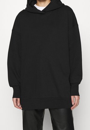 Person iført oversized sort hættetrøje og sorte læderbukser, stående mod en hvid baggrund.