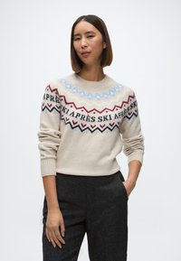 Kreemist lõngast kootud sviiter ümmarguse kaelusega, millel on sinised, punased ja mustad geomeetrilised mustrid. Koos tumehallide pükstega, mis rõhutavad mugavat lõiget.