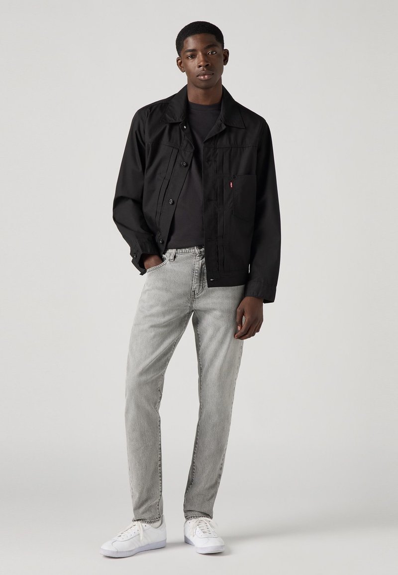 Zwart button-up jasje, grijze jeans en witte sneakers. Jasje heeft een borstzak en strakke lijnen; de jeans hebben een rechte snit en een vervaagde textuur.