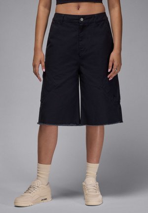 Schwarze Jeans-Shorts mit weitem Bein und fransigem Saum, ausgestattet mit zwei seitlichen Taschen und einem subtilen Logodetail. Dazu beigefügte beigefarbene Sneakers und Socken.