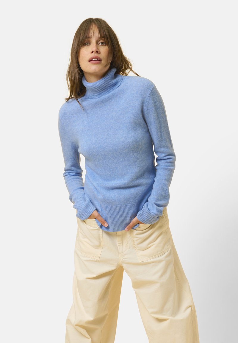 Maglione a collo alto blu chiaro realizzato in tessuto morbido. Presenta maniche lunghe, polsini e vita a coste. Abbinato a pantaloni larghi di colore chiaro.