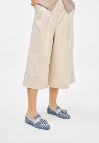 Mocassini intrecciati blu con una texture decorata e accenti blu chiaro, dotati di un dettaglio a fiocco e suola piatta. Abbinati a pantaloni beige a gamba larga.