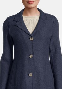 Marineblauer Wollmischungsoverall mit Reverskragen, fünf Knöpfen und zwei vorderen Taschen mit strukturierter Oberfläche. Ein Pullover blitzt darunter hervor.