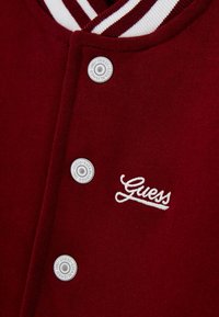 Burgunderfarbene Collegejacke mit weiß gestreiftem Kragen, Druckknöpfen und gesticktem "Guess"-Logo in Weiß. Weicher Stofftextur.