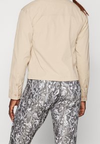 Giacca beige corta con polsini per bottoni, realizzata in un tessuto liscio. Abbinata a leggings grigi con motivo mimetico dal design aderente.
