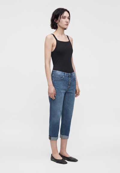 Femme aux cheveux courts foncés portant un haut noir sans manches, un jean bleu retroussé avec des boutons sur le devant et des chaussures plates noires, debout devant un fond blanc.