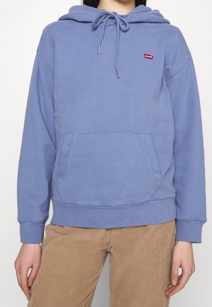 Person wearing a blue Levi's hoodie with front pocket and drawstring hood, paired with beige corduroy pants, against a white background.