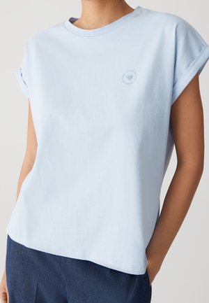 Person iført en lyseblå T-shirt med op rullede ærmer og et lille hjerte- og cirkeltekstlogo på brystet, kombineret med mørkeblå bukser.