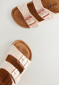 Glitzernde rosa Sandalen mit zwei verstellbaren silbernen Schnallen, weichen braunen Innensohlen und einer strukturierten Oberseite mit schimmernden Glitzerelementen.