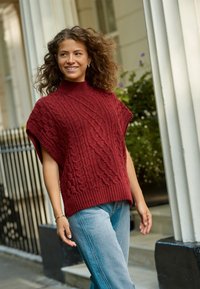 Pull en maille torsadée bordeaux avec col montant et manches courtes, associé à un jean droit bleu clair, sur fond d'un bâtiment.