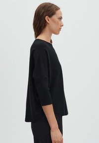 Femme en haut noir à manches trois-quarts et pantalon noir, debout de profil devant un fond gris clair uni.