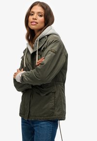 Olijfgroene parka-jack met een trekkoord in de taille, dubbele zakken en een grijze capuchon. Voorzien van een ritssluiting en een stoftextuur.