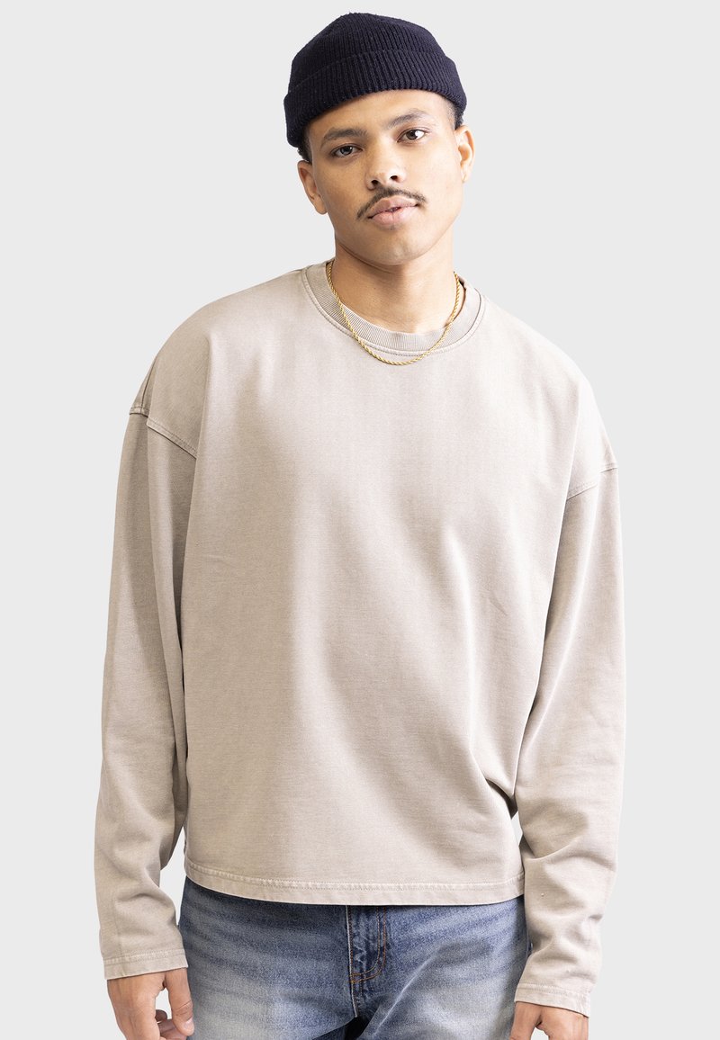 Beige langærmet sweatshirt med rund hals, afslappet pasform, rib strikkede manchetter og synlige sømme. Båret med blå jeans.