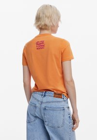 T-shirt in cotone arancione con maniche corte, caratterizzata da una grafica rosa sul retro. Indossata con jeans in denim azzurri chiari.