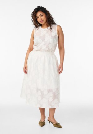 Kvinde med krøllet hår iført hvid blomsterprintet ærmeløs kjole og olivengrønne slingback-hæle, stående mod en hvid baggrund.