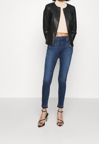 Giacca di pelle nera con un design aderente, top corto a righe beige, jeans skinny scuri strappati, e sandali neri con tacco alto e fasce.