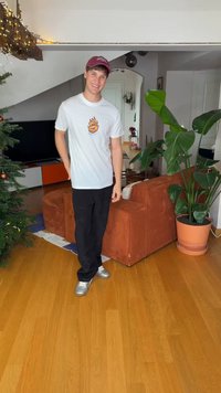 A person stands in a room, wearing a white t-shirt with a graphic design, black pants, and silver shoes. A brown corded sofa and green plant are visible.